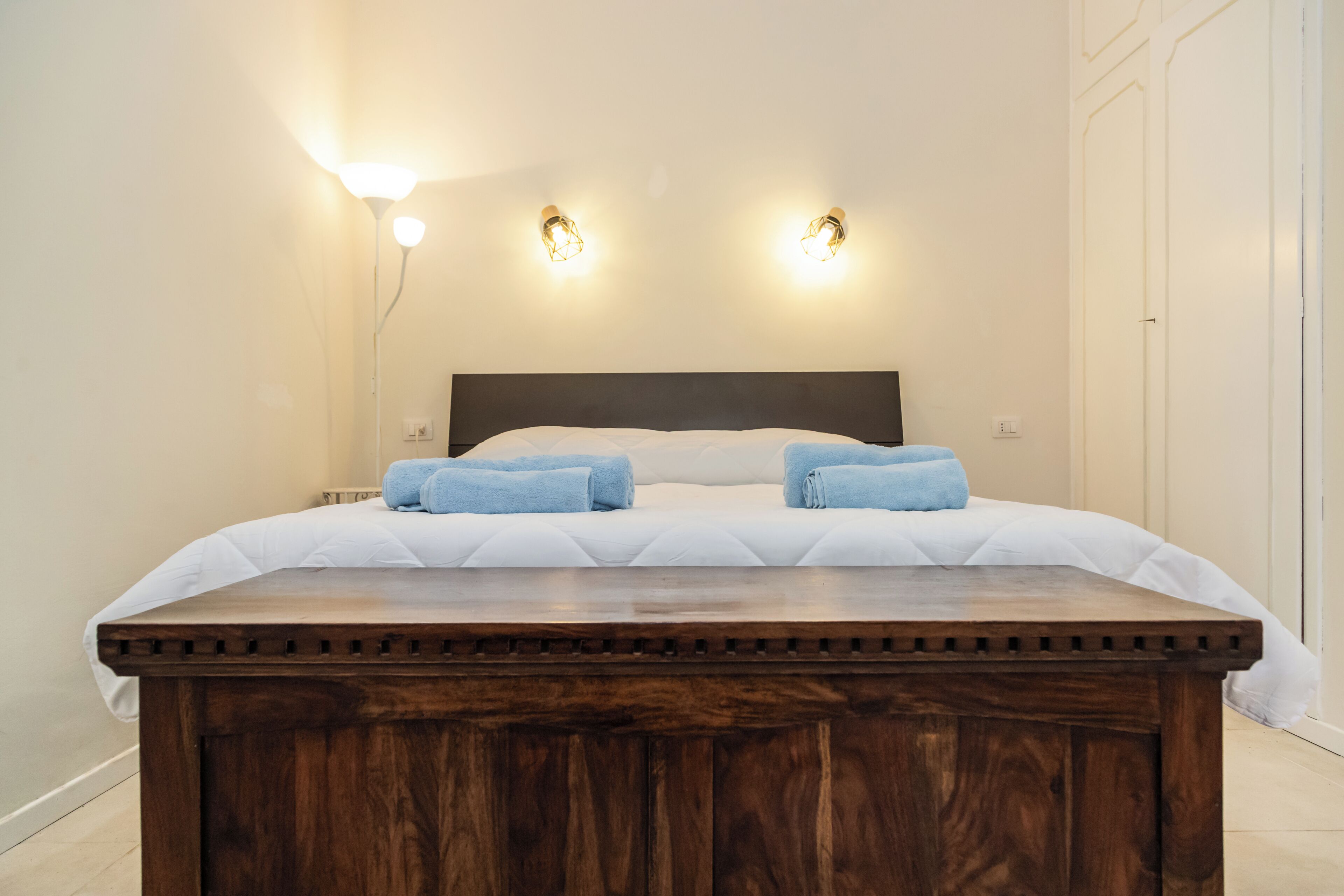 Una cama bien hecha con cabecera de madera, dos lámparas fijadas a la pared y una puerta blanca al fondo.