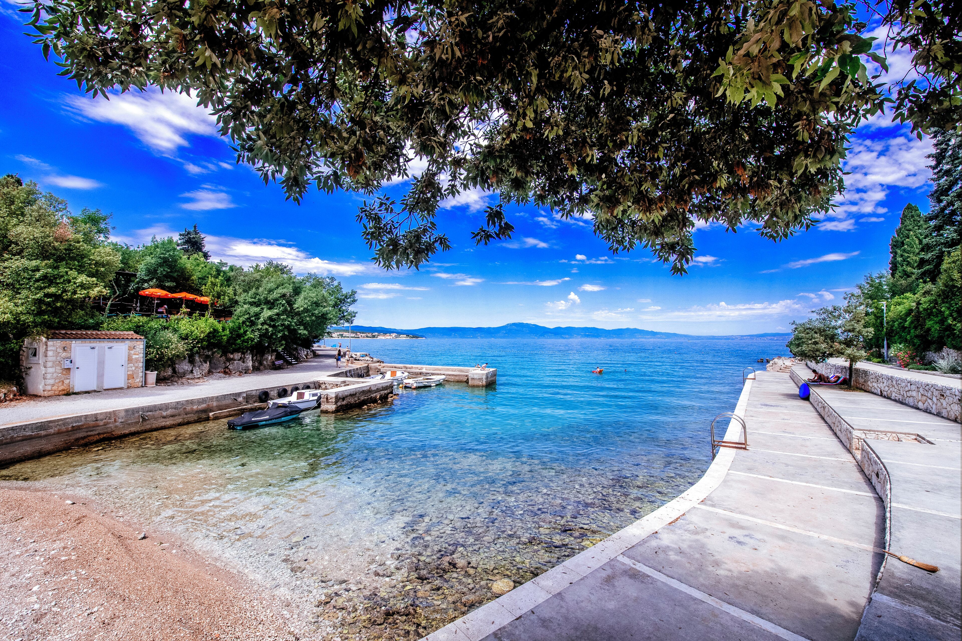 A coastal scene with a clear sky, calm water, a dock with boats, and a stone building.
