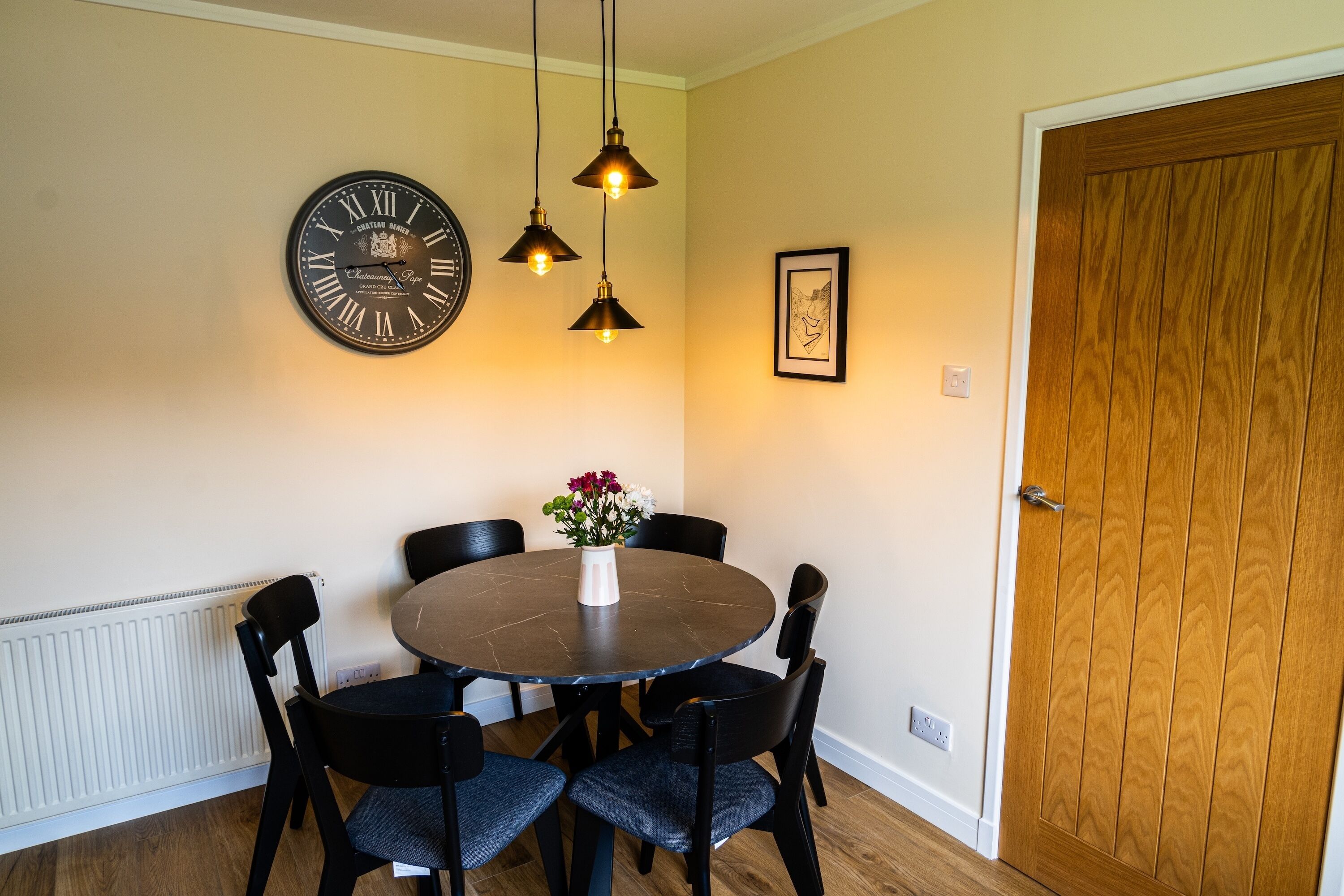 A dining area with a round table, six chairs, a wall clock, pendant lights, a framed picture, and a wooden door.