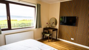 Une chambre à coucher comprenant un lit, un bureau, une chaise, un téléviseur et une fenêtre donnant sur un champ et des montagnes.