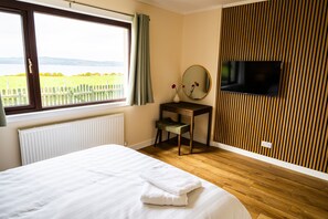 A bedroom with a bed, a desk, a chair, a TV, and a window with a view of a field and mountains. - Cozy Highland Cottage Escape (Inverness)
