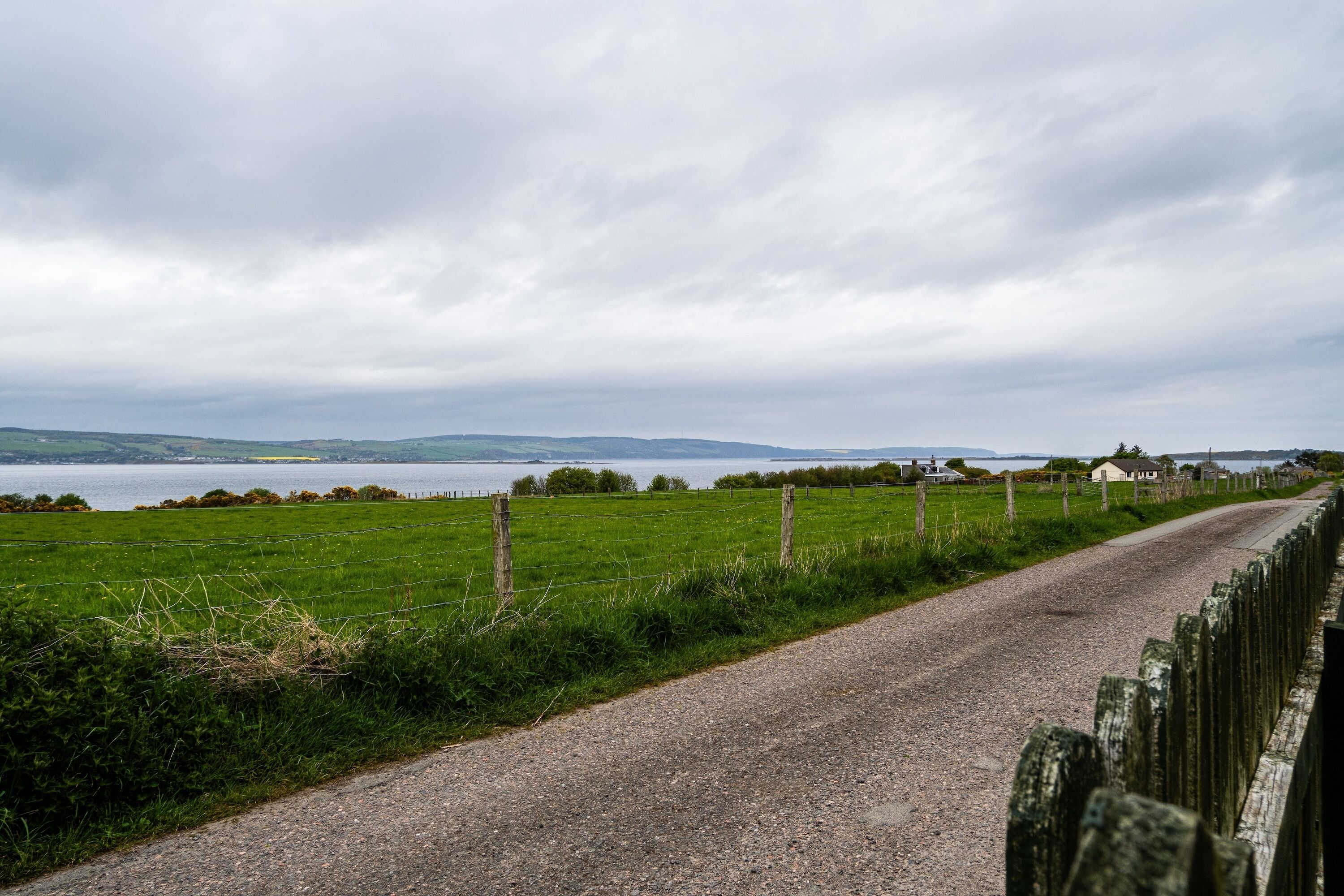 A gravel road with a fence on one side and a body of water in the distance.