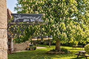 Ein Garten mit einem 'Bienvenue'-Schild, hängenden Laternen und Picknicktischen.