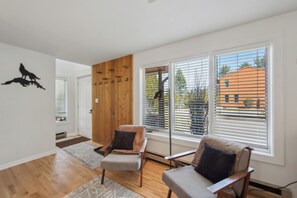 Ein modernes Wohnzimmer mit zwei Sesseln, einer Wand als Holzdekor, großen Fenstern mit Jalousien und Blick auf ein Gebäude.