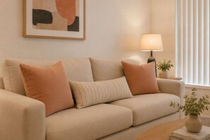 Ein beiges Sofa mit zwei Kissen, ein Beistelltisch mit Lampe, eine Vase mit Pflanze und ein gerahmtes abstraktes Gemälde an der Wand.