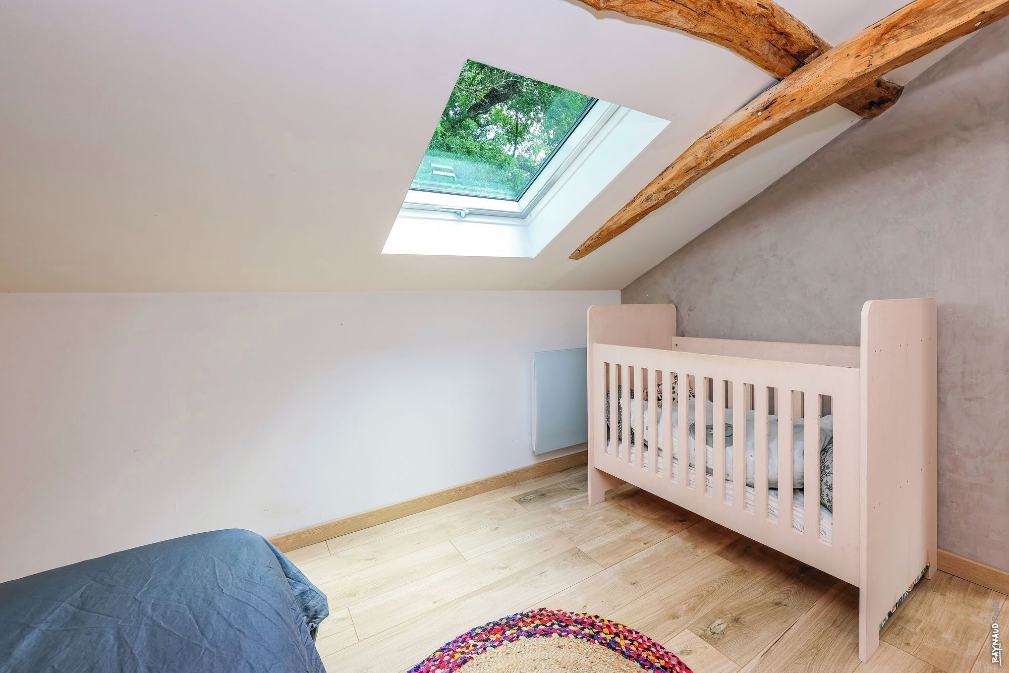 Een kamer met een schuine plafond, een wieg, een raam met uitzicht op groen en een kamerbreed tapijt met dessin.