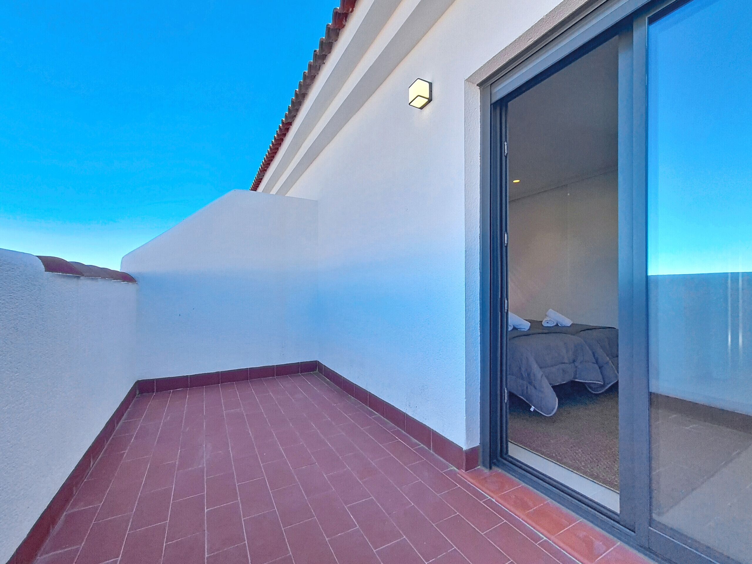Un balcone con tegole rosse, un letto con lenzuola bianche e una porta scorrevole in vetro che dà su una stanza.