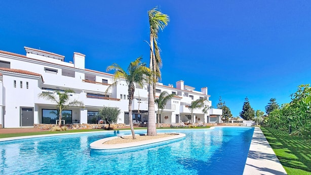 Una piscina con agua cristalina azul, rodeada por un edificio blanco con balcones y palmeras.