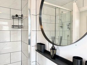 Een badkamer met witte marmertegels, een gebogen spiegel, een zwarte wastafel en een douchecabine van glas.