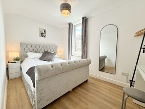 A bedroom with a tufted headboard, a bed with white linens, a bedside table with a lamp, a mirror, and a window with curtains. - Central Bristol 2-Bed w Roof Garden Wifi & Free Parking (Bristol)