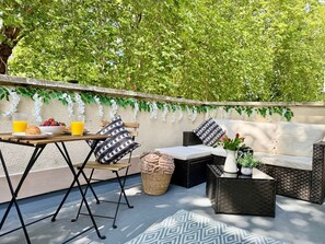 Een terras buiten met een tafel, stoelen, een bank en een rieten bijzettafeltje.