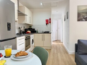 Een compacte keuken met een koelkast, fornuis, magnetron en een wasmachine. Er staat een eettafel met een bord eten en een glas sap.