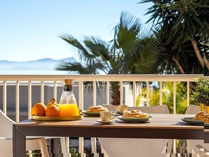 A table set with breakfast items, a glass of orange juice, and a bottle, with a view of palm trees and mountains in the background. - Chalet Mirador in Alcudia - Puerto de Alcudia (Alcúdia)