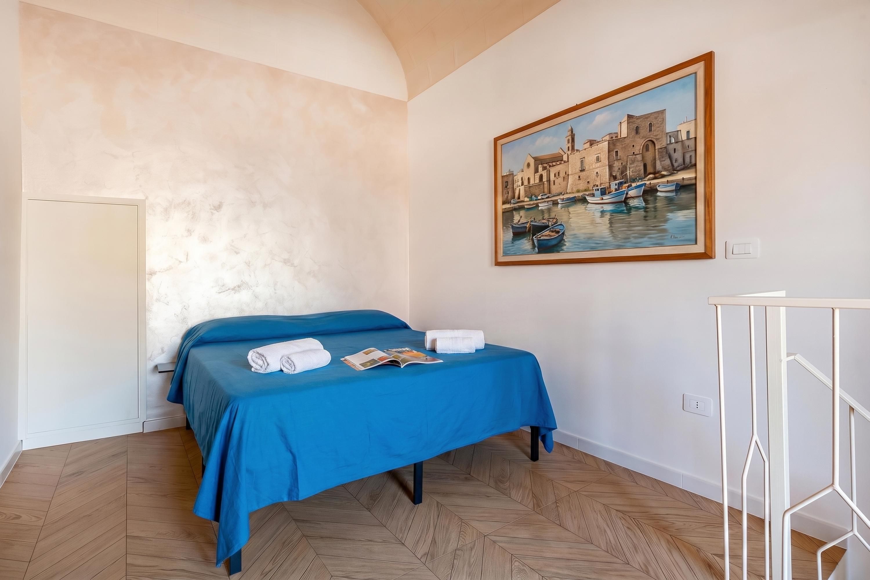 A hotel room with a bed, a painting of a coastal scene, and a wooden herringbone floor.