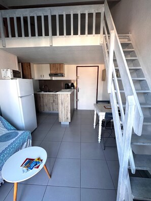 Una cocina compacta con microondas, refrigerador y armarios de madera. Una pequeña mesa de comedor con una silla se encuentra cerca de una escalera.