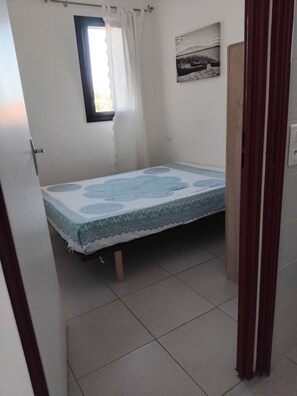 Une petite chambre avec un lit simple, une fenêtre, une porte et un tableau accroché au mur.