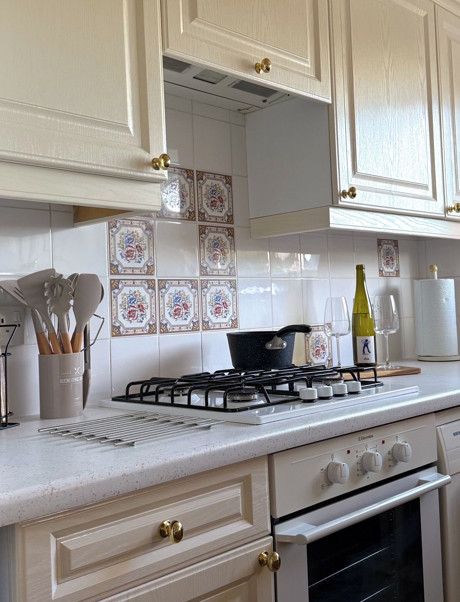 Une cuisine avec des meubles blancs, une cuisinière à gaz, une casserole, une bouteille de vin et un rouleau d’essuie-tout.