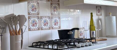 Une cuisine avec des meubles blancs, une cuisinière à gaz, une casserole, une bouteille de vin et un rouleau d’essuie-tout.