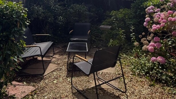 Un área de estar al aire libre con sillas y mesas, rodeada de follaje denso y plantas con flores.