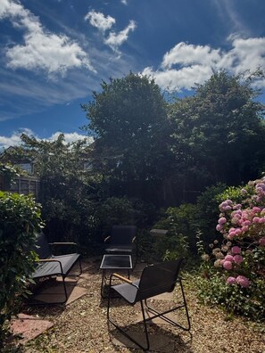 An outdoor seating area with chairs and tables, surrounded by dense foliage and flowering plants. - The Coastal Forest Retreat (Barton on Sea)