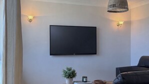 Una sala de estar con una televisión de pantalla plana montada en la pared, una silla de mimbre, un sofá, una mesa de centro de madera y una pequeña planta en maceta.