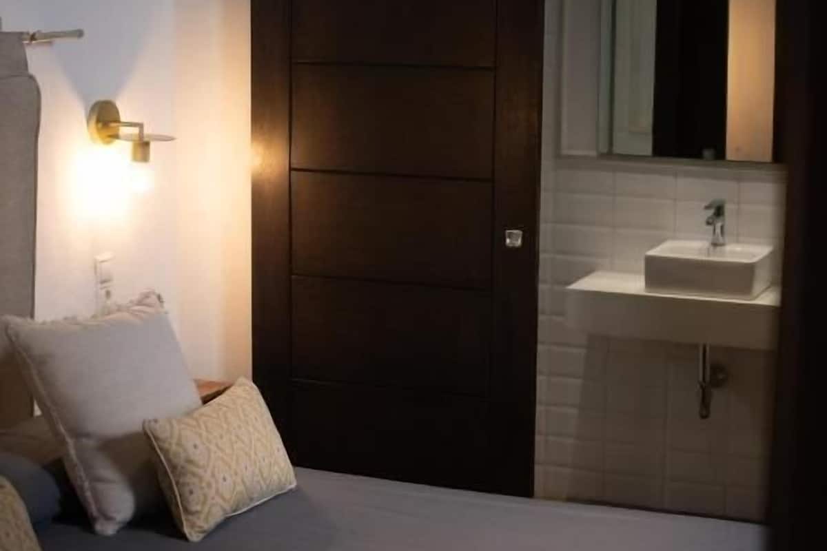 Un dormitorio con cama, mesita de noche, luz empotrada en la pared, una puerta y un baño con lavabo y espejo.