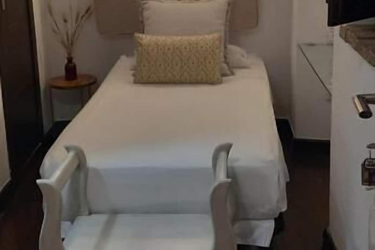 Un dormitorio pequeño y ordenado con cama, mesita de noche, silla y lámpara fijada a la pared.