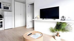 Un soggiorno moderno con un tavolino rotondo in legno, una TV a schermo piatto montata a parete, una piccola pianta in vaso e un angolo cucina con mobili e elettrodomestici bianchi.