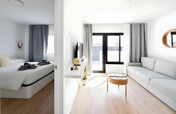 Ein modernes Hotelzimmer mit Bett, Sofa, Fernseher, kleinem Tisch und einem Balkon mit Vorhängen.
