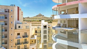 A multi-story residential building with balconies, a view of a hill, and construction materials visible. - Modern Seaside Apart in Marsalforn - 5E, Marsalforn Iz-Zebbug Gozo, Malta (Marsalforn Iz-Zebbug Gozo)