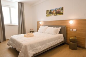 Een slaapkamer met een bed, nachtkastje, wandlamp, raam met gordijnen en een schilderij aan de muur.