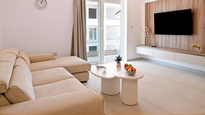 A modern living room with a sofa, a flat-screen TV, a small table with a fruit bowl, and a sliding glass door. - Modern Seaside Apart in Marsalforn - 5E, Marsalforn Iz-Zebbug Gozo, Malta (Marsalforn Iz-Zebbug Gozo)