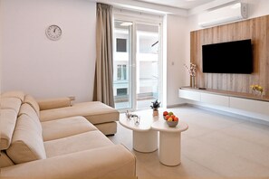 Ein modernes Wohnzimmer mit einer Couch, einem Flachbildfernseher, einem kleinen Tisch mit Obstschale und einer Schiebetür aus Glas.