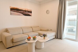 Een moderne woonkamer met een beige bank, twee witte bijzettafels met fruitschaaltjes, een klok en een stadsgezicht schilderij.