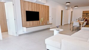 A modern living room with a flat-screen TV, white sofas, a dining area, and a kitchen in the background. - Modern Seaside Apart in Marsalforn - 6A, Marsalforn Iz-Zebbug Gozo, Malta (Marsalforn Iz-Zebbug Gozo)