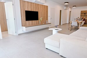 A modern living room with a flat-screen TV, white sofas, a dining area, and a kitchen in the background. - Modern Seaside Apart in Marsalforn - 6A, Marsalforn Iz-Zebbug Gozo, Malta (Marsalforn Iz-Zebbug Gozo)