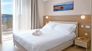 A hotel room with a large bed, a nightstand, a window with curtains, and a painting on the wall. - Modern Seaside Apart in Marsalforn - 6A, Marsalforn Iz-Zebbug Gozo, Malta (Marsalforn Iz-Zebbug Gozo)