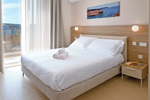 A hotel room with a large bed, a nightstand, a window with curtains, and a painting on the wall. - Modern Seaside Apart in Marsalforn - 6A, Marsalforn Iz-Zebbug Gozo, Malta (Marsalforn Iz-Zebbug Gozo)