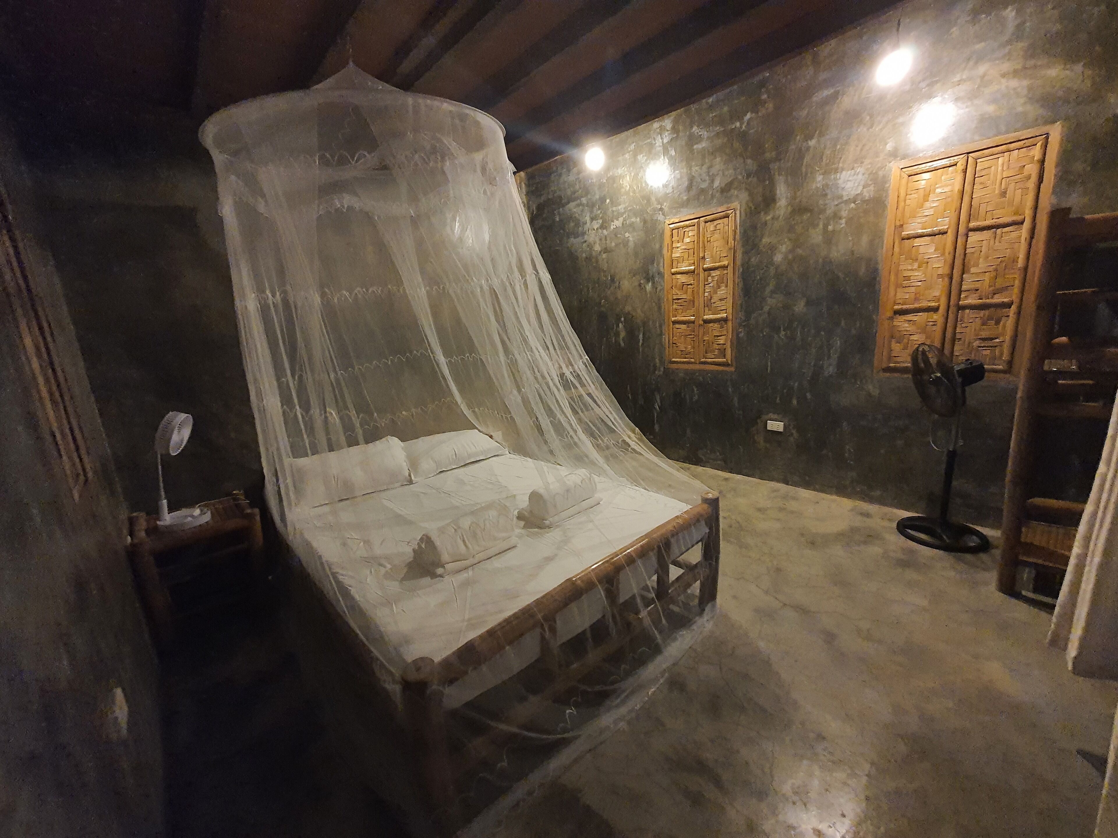 Una habitación con una cama bajo una estructura tipo dosel, un armario de madera, un ventilador de pie y una mesita con una lámpara.