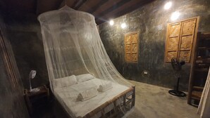 A room with a bed under a canopy, a wooden wardrobe, a standing fan, and a small table with a lamp. - Coleto Siargao (Rodriguez)