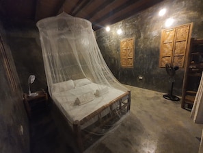Una habitación con una cama bajo una estructura tipo dosel, un armario de madera, un ventilador de pie y una mesita con una lámpara.