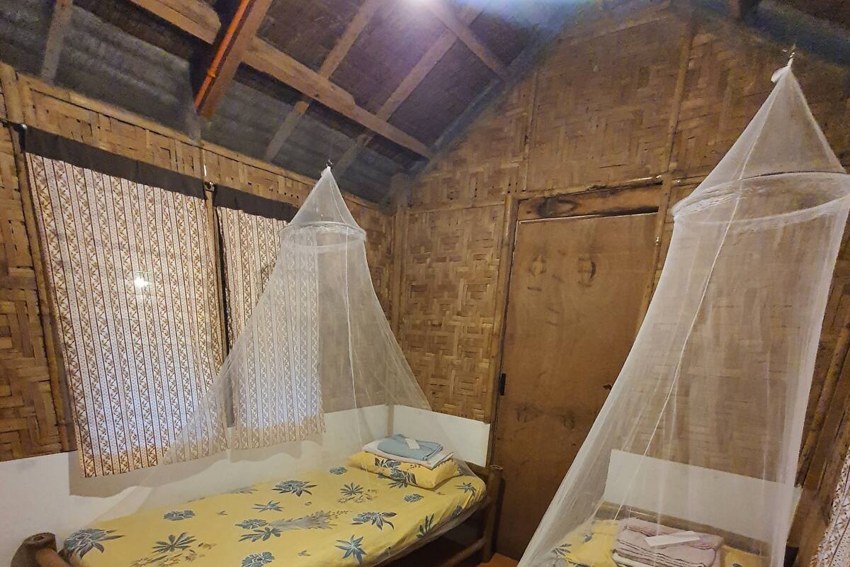 Zwei hölzerne Betten mit Moskitonetzen, eine gewebte Wand und eine Holzbalkendecke.