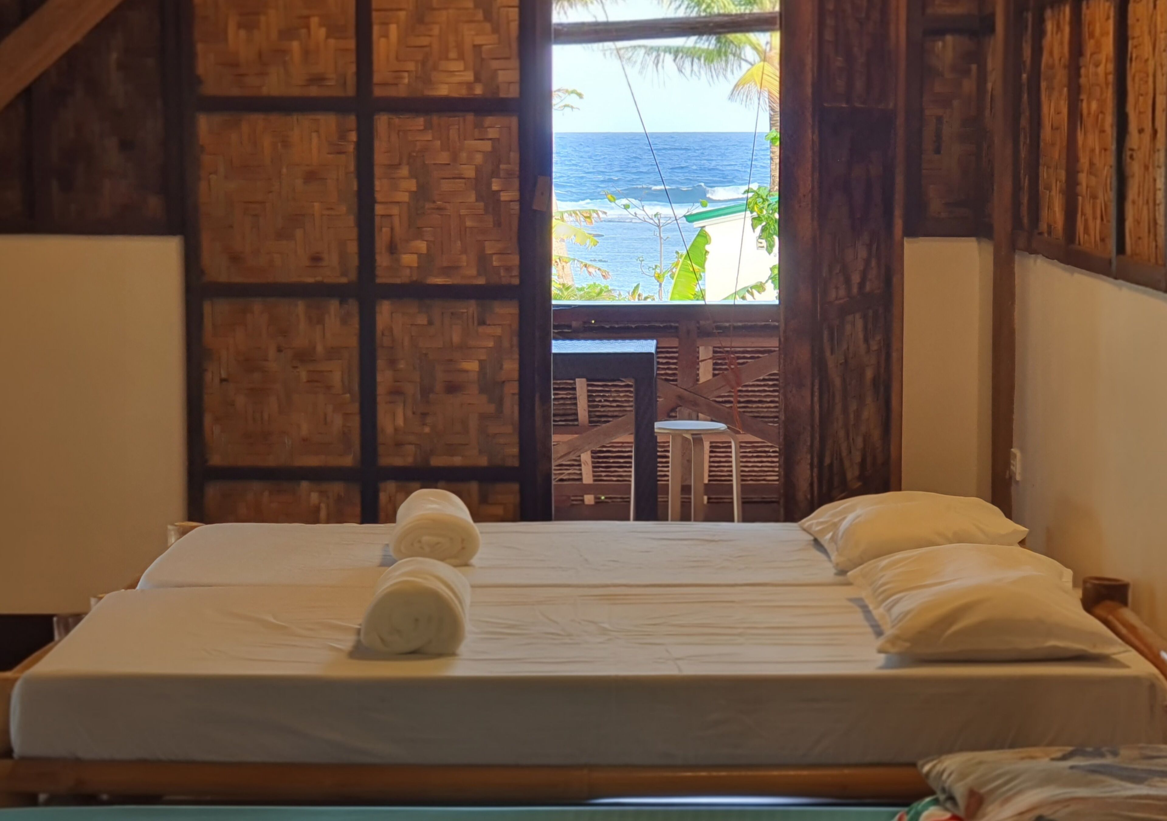 Una cama con sábanas blancas y una toalla, un muro de madera y vista al océano a través de una puerta abierta.