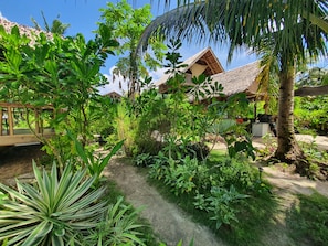 Un jardín tropical con una estructura de techo de paja, abundante vegetación y cielo despejado.