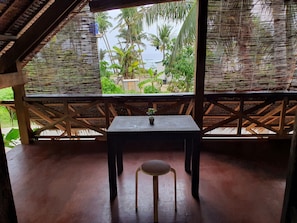Un espacio de estar al aire libre techado, con una mesa, una silla, vigas de madera y vistas a zonas verdes y edificios.