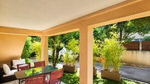 A covered patio with a glass table and red chairs, surrounded by potted plants and a view of a street. - Chez Aurel et Ju (Tournon-sur-Rhône)