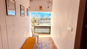 A hallway with a bench, framed pictures on the wall, and a view of a building with mountains in the background. - Milam Hostel (Leh)