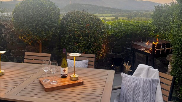 An outdoor patio with a table set for two, a bottle of wine, and glasses. There is a view of distant hills and a stone wall. - The Stirling Lookout | Panoramic Terrace Views | Steps from Stirling Castle (Stirling)