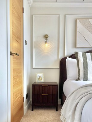 A bedroom with a wooden door, a bedside table with a picture frame, a wall-mounted light, and a bed with pillows.