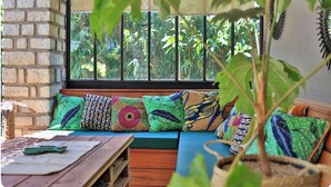 Un rincón acogedor con un banco y cojines, una mesa de madera y vistas al exterior a través de una ventana que muestra un muro de ladrillo y vegetación.
